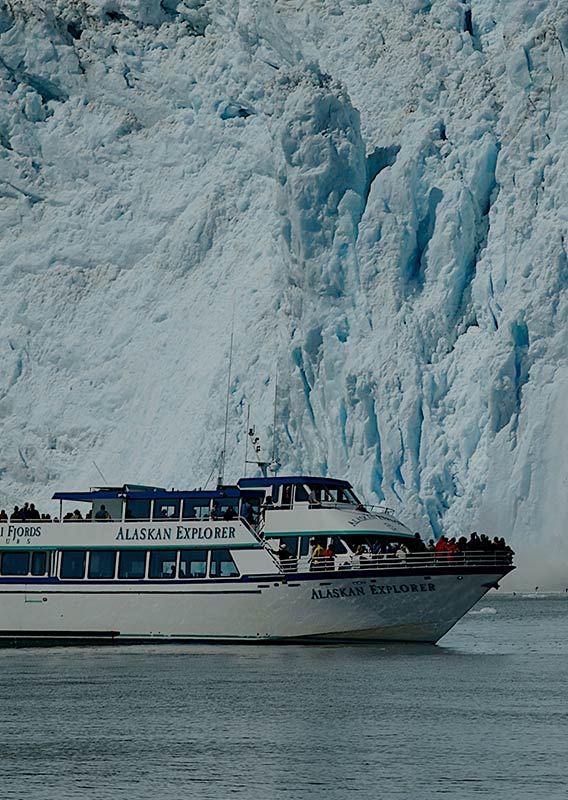 The Alaskan Explorer boat cruises through the water next to a large iceberg.