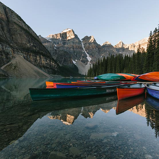A series of canoes docked on a clear lake below tall snow-capped mountains.