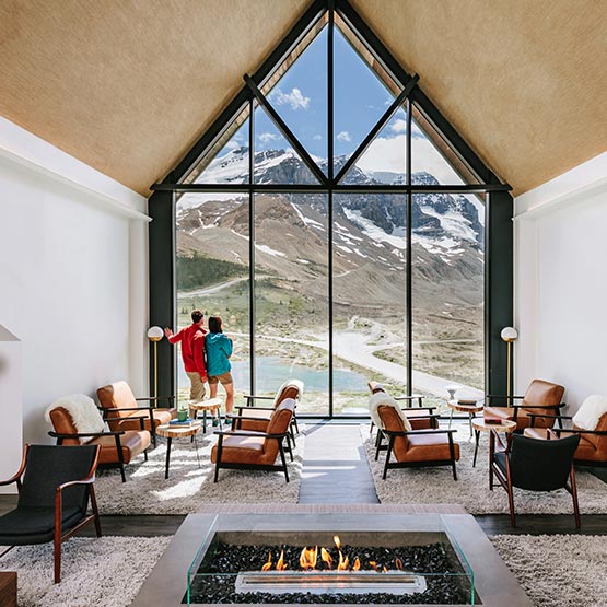 Two people stand at a large window in a lounge overlooking a high alpine landscape.