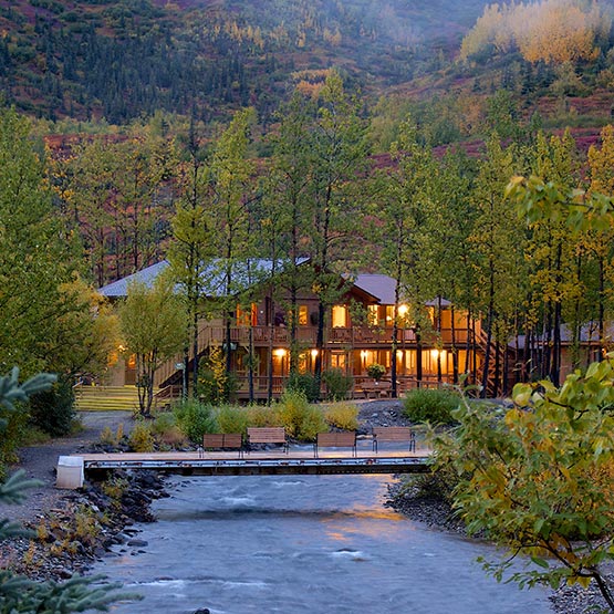 A wooden lodge building lit up at dusk, alongside a river and forest.
